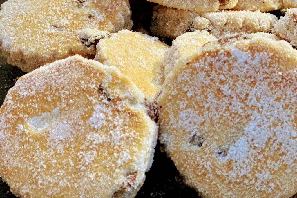 Traditional Welsh cakes stacked on a rustic plate beside a steaming cup of tea, softly lit by afternoon sunlight through a kitchen window—sugar-dusted and golden, evoking a cosy, homely tea-time scene in the heart of Wales.